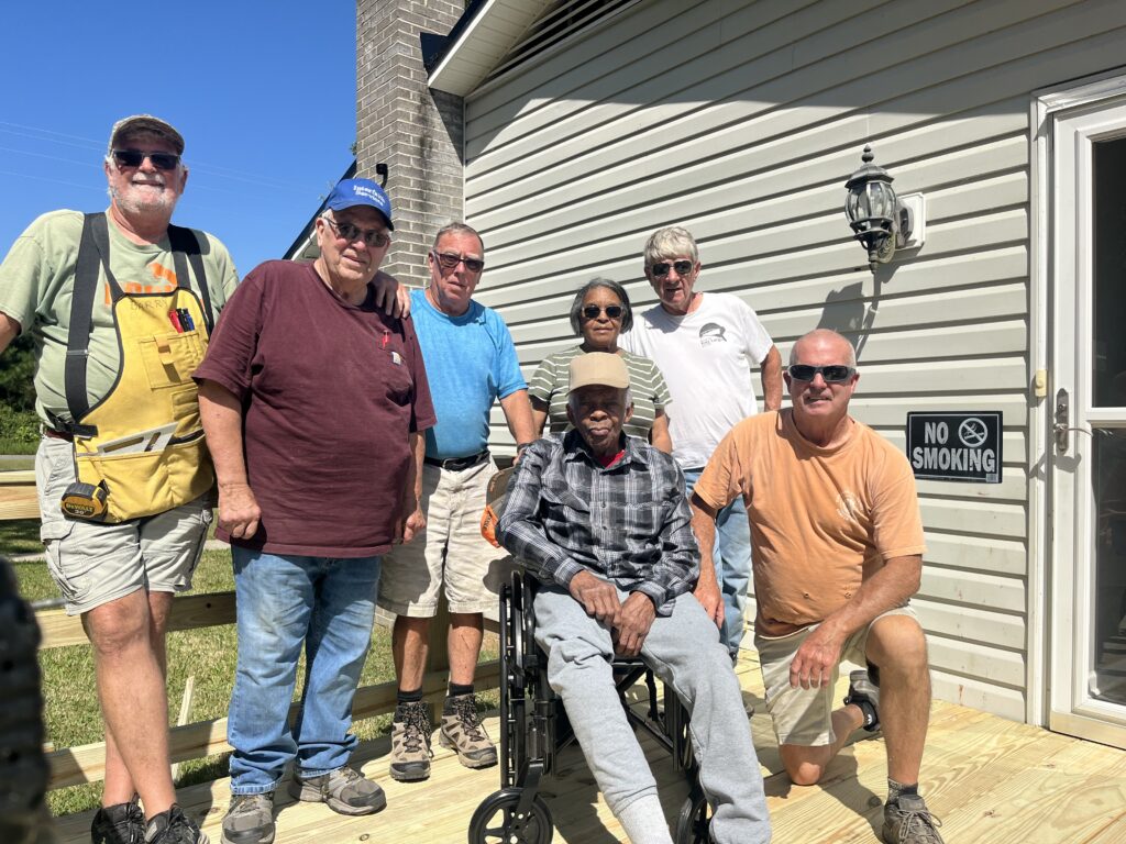 A group of Interfaith volunteers including men and women pose on a newly built ramp with a person in their wheelchair. The sky behind them is bright blue.